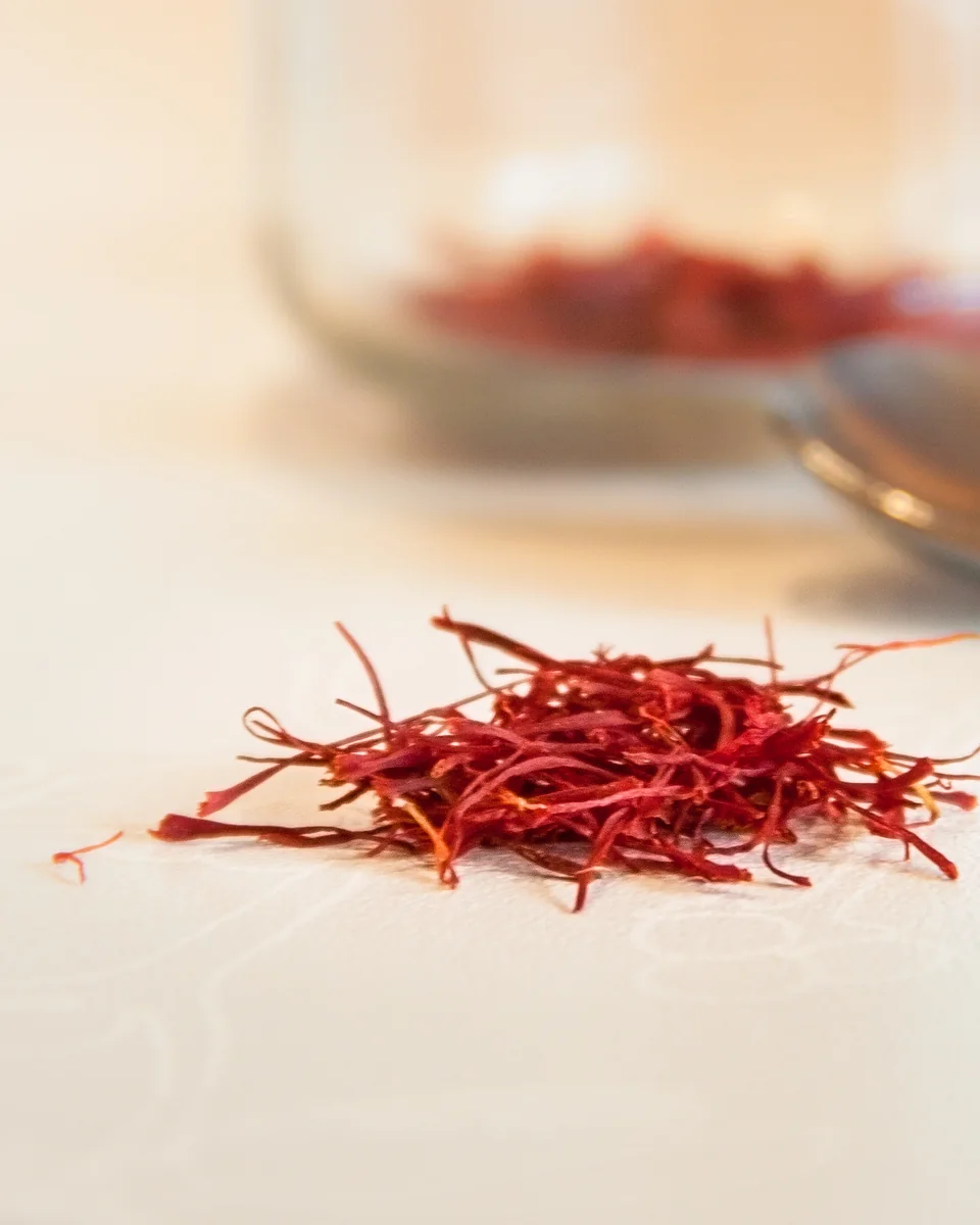 A small pile of saffron threads on a pale surface with a softly blurred spoon and glass jar behind it.